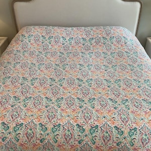 May include: A bed quilt featuring a repeating damask-style pattern in shades of teal, coral, and soft pink on a white background, draped over a bed with a light-colored, arched, upholstered headboard with a wooden frame.