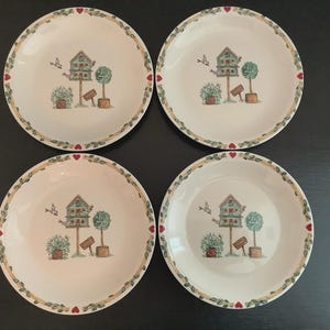 May include: A set of four round white ceramic plates featuring a central illustration of a birdhouse on a post, a small tree, and a bird in flight. The rim is decorated with a green leaf vine pattern interspersed with small red heart icons.