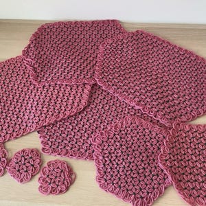 May include: A collection of crocheted mats and coasters in varying sizes, featuring a repeating floral-like pattern in dusty rose pink yarn with dark green centers. The set includes several rectangular and circular pieces with scalloped edges.