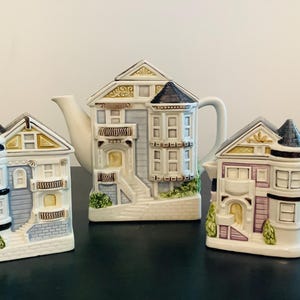 以下が含まれることがあります： ビクトリア様式の家を模した陶器製のティーセット。ティーポット、クリーマー、シュガーボウルの3点セット。ティーポットとシュガーボウルは水色、クリーマーは薄紫色で、それぞれ濃い色の屋根と装飾が施されている。