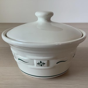 May include: A cream-colored ceramic bowl with a matching lid. The bowl features a decorative band around the center with dark green geometric floral motifs and curved lines. A thin dark green horizontal line circles the base of the bowl.
