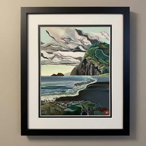 May include: A framed landscape painting depicts a coastal scene with a dark sand beach, blue ocean waves, and a large green cliff under a sky filled with layered gray and white clouds. The artwork is enclosed in a black frame with a white mat.