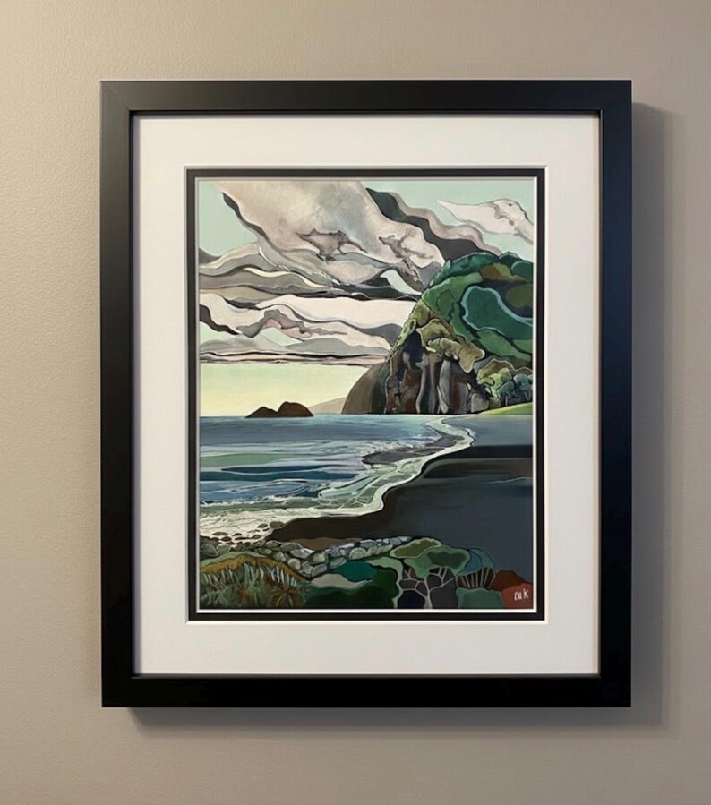 May include: A framed landscape painting depicts a coastal scene with a dark sand beach, blue ocean waves, and a large green cliff under a sky filled with layered gray and white clouds. The artwork is enclosed in a black frame with a white mat.
