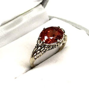 May include: A silver-toned ring featuring an oval-cut, deep orange gemstone set in a prong setting. The band of the ring displays an intricate, openwork filigree design that extends from the base of the gemstone setting down the sides of the band.