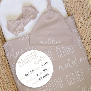 May include: A newborn baby set in a woven basket, including a blanket, hat, and bow. The blanket is taupe with the name "Madeline Claire" in a repeating pattern. A white circle displays the baby's birth details: date, time, weight, and length.