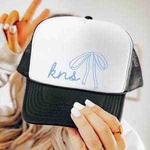May include: A black and white trucker hat with a white front panel. The front panel has a blue bow and the text "kns" in blue.