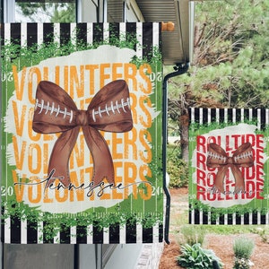 May include: Two garden flags with a football theme. The larger flag is green, black, and white with the words "Volunteers" and "Tennessee" in yellow. The smaller flag is black, white, and red with the words "Roll Tide" in red. Both flags feature a brown football-shaped bow.