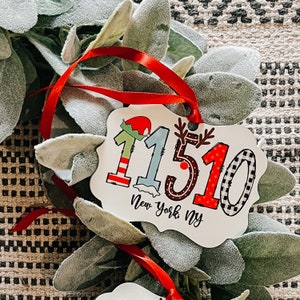 May include: A white ornament with the number "11510" in festive colors and patterns, including red, green, and black, with the text "New York, NY". The ornament is hanging with a red ribbon, surrounded by green foliage.
