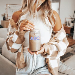 May include: A woman wearing a white tank top and a brown and white checkered cardigan holds a glass jar with a lid. The jar is filled with a brown liquid and has the letters "ABC" printed on the front in pink and blue.