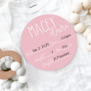 May include: A pink circular birth announcement with the name "Macey Claire" in white script. The announcement includes the date, time, weight (7 lbs 14.6 oz), and length (21.75 inches). A wooden baby rattle and a white onesie are also in the image.