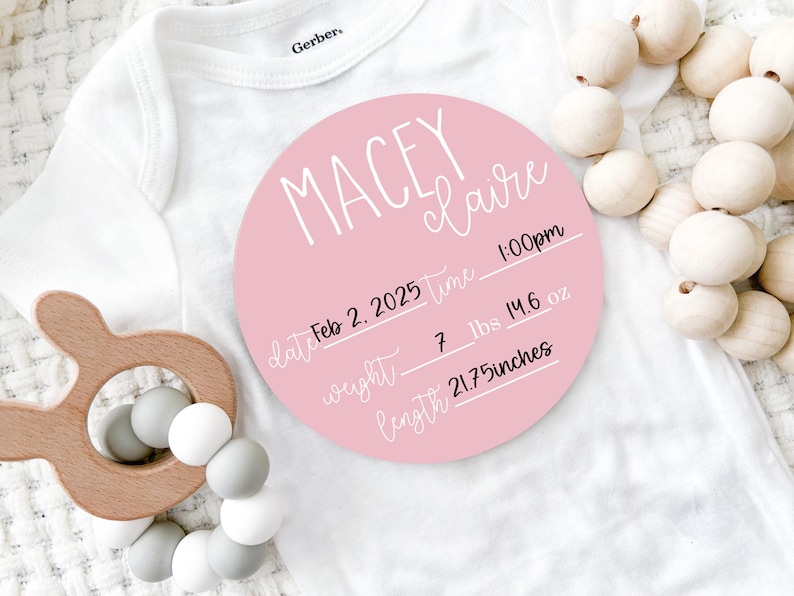 May include: A pink circular birth announcement with the name "Macey Claire" in white script. The announcement includes the date, time, weight (7 lbs 14.6 oz), and length (21.75 inches). A wooden baby rattle and a white onesie are also in the image.