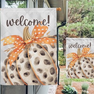 May include: A decorative garden flag featuring a watercolor pumpkin design with a polka-dot orange bow and the word "welcome!". The pumpkin has brown spots and is set against a white background with a diamond pattern. A smaller version is also displayed.