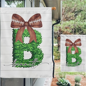 May include: A white garden flag featuring a green letter "B" crafted from leaves and a brown football-stitched bow. The name "bennett" is written in cursive beneath the letter.