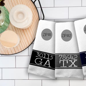 May include: Three white hand towels with black, grey, and navy blue bands featuring zip codes and state abbreviations. The towels are labelled ZIP #1, ZIP #2, and ZIP #3. The towels are displayed on a white tiled surface.