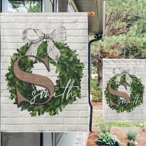 May include: A white brick background with a green wreath featuring a brown burlap letter "S" at its centre. A white and black striped bow sits atop the wreath. The text "Smith" is written in cursive below the wreath.