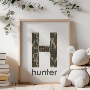 Pode incluir: Uma impressão emoldurada com uma letra H de padrão de camuflagem e a palavra "hunter" abaixo. A impressão está sobre um fundo branco e está emoldurada em uma moldura de madeira clara.