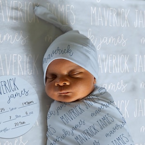 May include: A newborn baby wrapped in a swaddle blanket and wearing a matching hat, both with the name "Maverick James" printed on them. A circular sign displays the baby's birth details: date, time, weight (7 lbs, 6 oz), and length (20.5 inches).