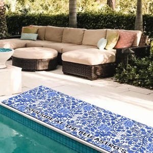 May include: A blue and white patterned poolside mat with the text "Poolside at the Belcastro's" printed on it. The mat is placed on the edge of a swimming pool.