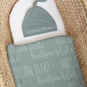 May include: A baby blanket and matching cap in a woven basket. The blanket and cap are a muted teal color with the name "hudson blake" printed in a repeating pattern. The cap is knotted at the top.