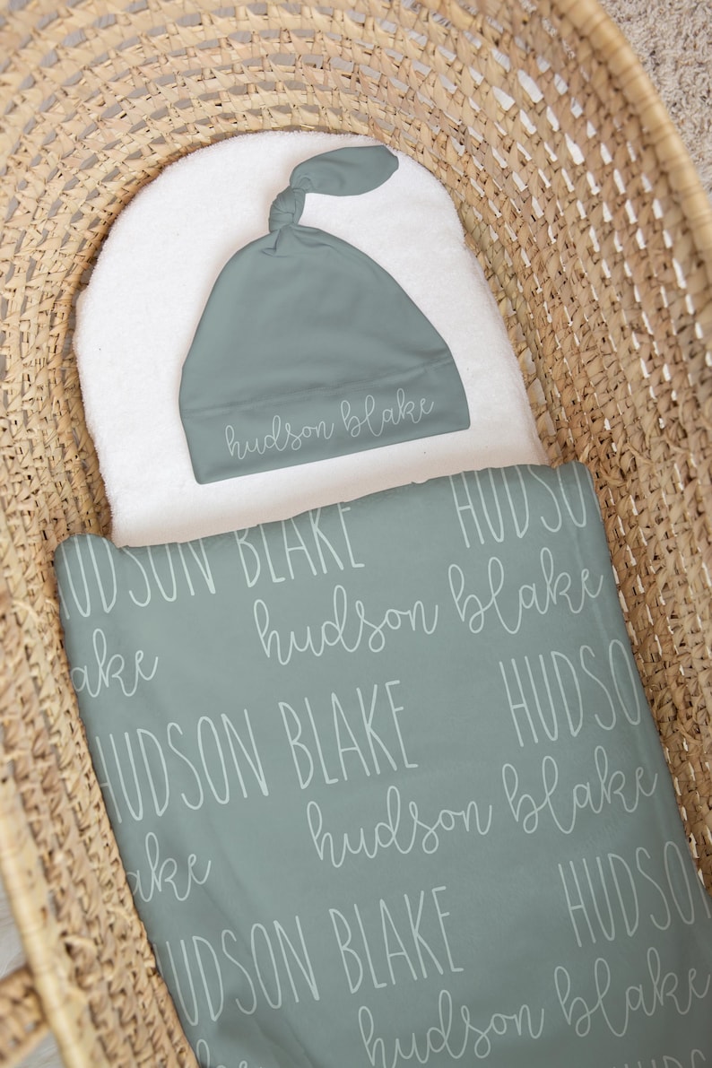 May include: A baby blanket and matching cap in a woven basket. The blanket and cap are a muted teal color with the name "hudson blake" printed in a repeating pattern. The cap is knotted at the top.