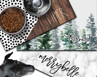 Pode incluir: Três tapetes para tigelas de cães com designs diferentes. O primeiro tapete tem um padrão de bolinhas pretas e brancas. O segundo tapete tem um design de aquarela de pinheiros com o nome "Bernard" escrito em itálico. O terceiro tapete tem um design de mármore branco com o nome "Merrybelle" escrito em itálico.