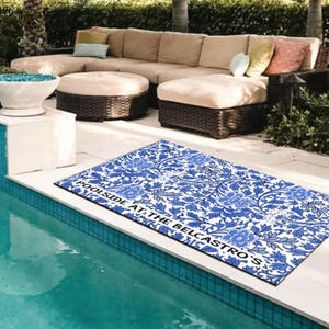 May include: A blue and white patterned doormat with the text "Poolside at the Belcastro's" in black lettering. The doormat is placed on a white tiled surface next to a swimming pool.