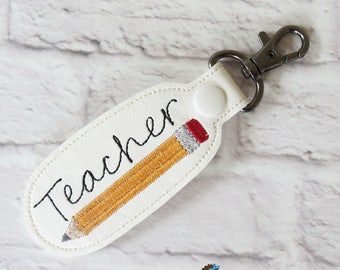 May include: White keychain with the word "Teacher" in black script and a yellow pencil design. The pencil has a red eraser and a gray tip. The keychain has a metal clasp.