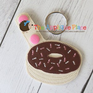 May include: A cream-colored donut-shaped keychain with a brown chocolate frosting and pink and white sprinkles. The keychain has a metal ring and a cream-colored strap with a cartoon hedgehog and pink circles. The text "The Applique Place" is visible.