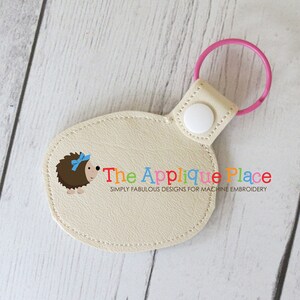 May include: A beige, oval-shaped keychain with a pink ring and white snap closure. It features a cartoon hedgehog with a blue bow and the text "The Applique Place" in colorful letters. The design is for machine embroidery.