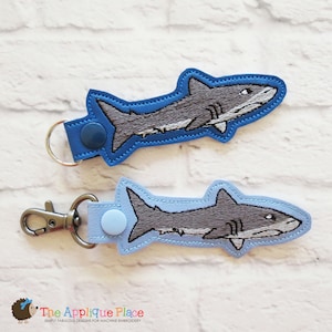 May include: Two shark-shaped keychains. The sharks are gray with white accents, outlined in blue, and attached to blue or light blue straps. One keychain has a silver keyring, the other a silver clasp. The Applique Place logo is in the bottom left.