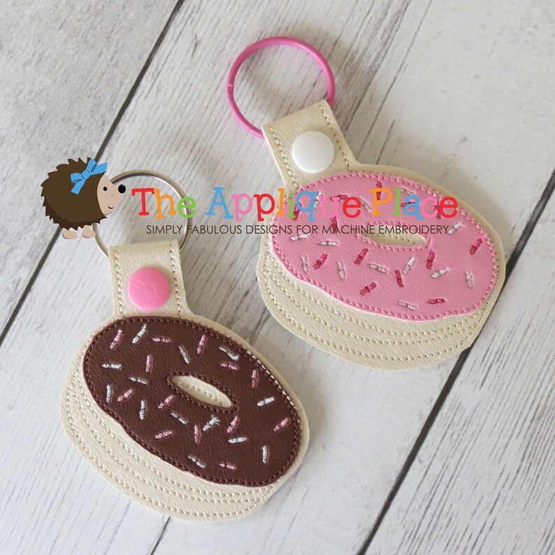 May include: Two donut-shaped keychains. One has a chocolate frosting design with brown sprinkles, and the other has a pink frosting design with white sprinkles. Both keychains have a beige base, pink or white button, and a metal ring.
