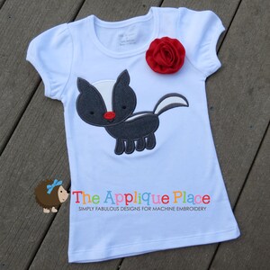 Puede incluir: Camiseta blanca de manga corta con un dise&ntilde;o de mofeta negro y blanco bordado. La camiseta tiene una flor de tela roja en la parte delantera. El texto "The Applique Place" est&aacute; impreso en la parte inferior de la camiseta.