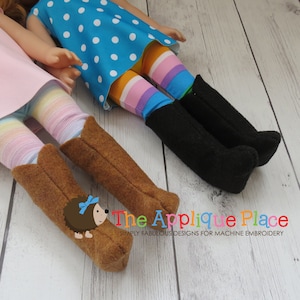 Pu&ograve; includere: Due bambole che indossano abiti e stivali diversi. Una bambola indossa un vestito blu a pois con leggings a righe arcobaleno e stivali neri. L'altra bambola indossa un vestito rosa con leggings a righe arcobaleno e stivali da cowboy marroni. Le bambole sono sdraiate su una superficie di legno bianco. Il testo "The Applique Place" &egrave; visibile nell'angolo in basso a destra dell'immagine.