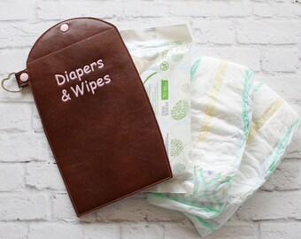 May include: A brown faux leather diaper and wipes pouch with the words "Diapers & Wipes" embroidered in pink. The pouch is open, revealing a package of wipes and two diapers.