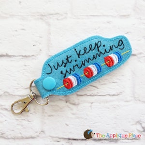 May include: A turquoise keychain with the phrase "Just keep swimming" embroidered in black. The keychain features three red, white, and blue swimming lane markers. It has a silver clasp and a blue button.