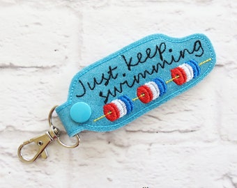 May include: A turquoise keychain with the phrase "Just keep swimming" embroidered in black. The keychain features three red, white, and blue swimming lane markers. It has a silver clasp and a blue button.