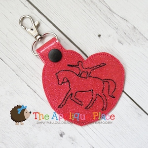 Op de afbeelding: Een roze glitter hartvormige sleutelhanger met een zwarte knop en een zilveren sleutelring. De sleutelhanger heeft een zwart geborduurd ontwerp van een persoon die een voltige-oefening op een paard uitvoert.