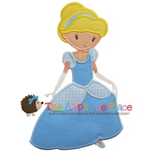 Op de afbeelding: Geborduurde applicatie van een blauw geklede Assepoester met een gele knot en witte handschoenen. Ze houdt een witte toverstaf vast en heeft een klein bruin egeltje met een blauwe strik naast haar. De tekst "The Applique Place" staat onder haar.