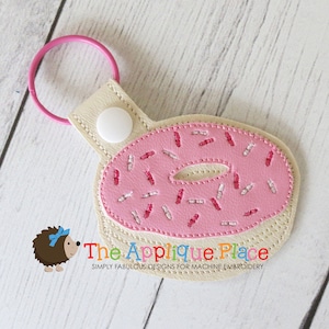 May include: A pink donut-shaped keychain with white and pink sprinkles. The keychain has a light pink ring and a beige backing with a white snap closure. The text "The Applique Place" is visible.