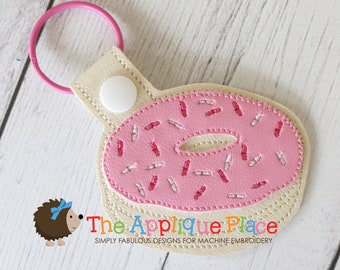 Può includere: Un portachiavi a forma di ciambella rosa con zuccherini bianchi e rosa. Il portachiavi ha un anello rosa chiaro e un retro beige con una chiusura a scatto bianca. Il testo "The Applique Place" è visibile.