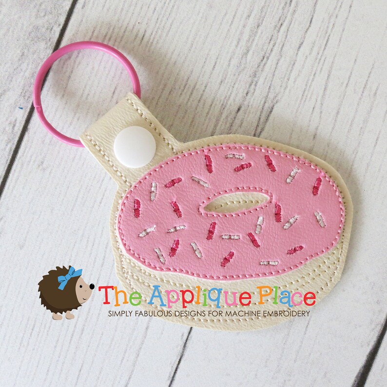 May include: A pink donut-shaped keychain with white and pink sprinkles. The keychain has a light pink ring and a beige backing with a white snap closure. The text "The Applique Place" is visible.