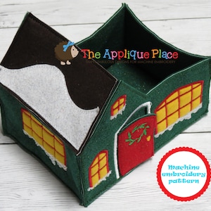May include: A green felt house-shaped storage box with a brown roof and white snow accents. The house features yellow window details, a red door with a green wreath, and the text "The Applique Place". A red circle with the text "Machine embroidery pattern" is also visible.