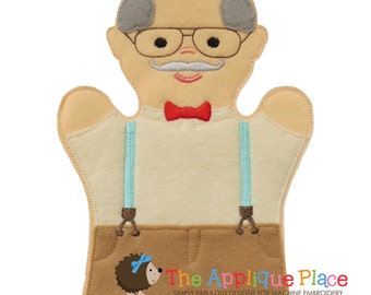 May include: A felt puppet of a person with grey hair, glasses, a moustache, a red bow tie, and blue braces. The puppet is wearing a beige shirt and brown trousers. The text "The Applique Place" is on the bottom of the puppet.