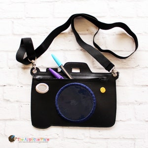 May include: Black camera-shaped crossbody bag with a black strap. The bag has a navy blue circle with blue stitching, a grey oval, and a gold button. Two pens are visible inside the bag. The bag is designed to look like a camera.