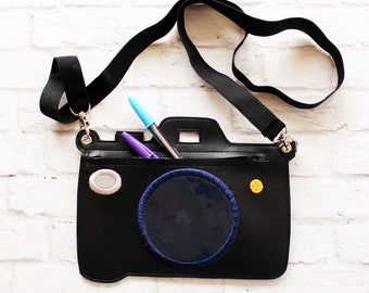 May include: Black camera-shaped crossbody bag with a black strap. The bag features a navy blue circle with blue stitching, a gray oval, and a gold button. Two pens are visible inside the bag. The bag is designed to look like a camera.