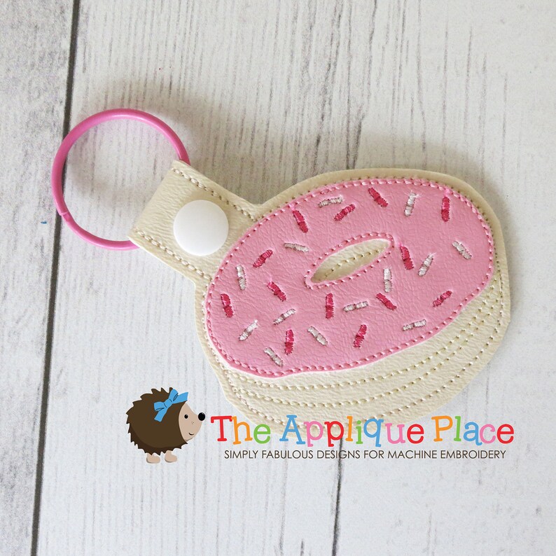 May include: A pink donut-shaped keychain with pink sprinkles and a light beige base. The keychain has a pink ring and a white button. The Applique Place logo is visible in the bottom right corner.