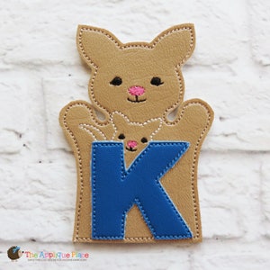 Puede incluir: Una marioneta de canguro marrón con un bebé canguro en su bolsa. La marioneta tiene una letra "K" azul cosida en la parte delantera. La marioneta tiene ojos negros y una nariz rosa. El logotipo de The Applique Place está en la esquina inferior izquierda.