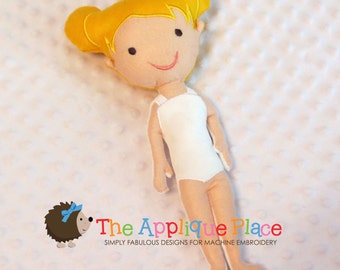 May include: A handmade felt doll with yellow hair styled in two buns. The doll has a white leotard and tan limbs. The face features black eyes and a stitched pink smile. The doll is lying on a textured, light pink surface. The Applique Place logo is in the bottom right.