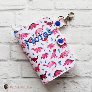 May include: A white notebook cover featuring a pattern of pink dinosaurs and the word "Notes" embroidered in blue. The cover has a snap closure with blue buttons and a silver-coloured clip. A pen is inserted into the side.