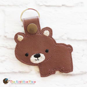 May include: A brown bear keychain with a white face and black eyes. The keychain is made of faux leather and has a metal ring.